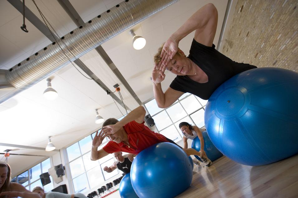 Gruppenfitness im Studio: Teilnehmer üben auf blauen Gymnastikbällen mit Blick zur Fensterfront.